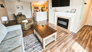 A modern living room with a sofa, a coffee table, and a fireplace. There is a kitchen area with a microwave and a TV mounted above the fireplace. - 2br/2ba Full Condo w/ Pool & Gym, Sleeps 6 (Gulf Shores)