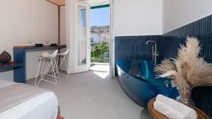 A modern hotel room with a bed, a small dining area with chairs, a blue bathtub, and a view of the outside. - Suite Pina Ocean in Sorrento (Sorrento)