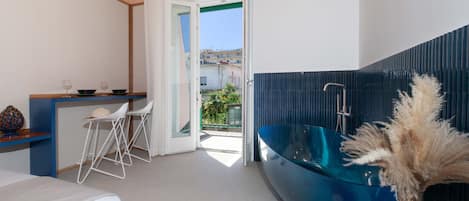 A modern hotel room with a bed, a small dining area with chairs, a blue bathtub, and a view of the outside.