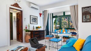 A living room with a laptop on a table, a sofa with cushions, a dining area with a table and chairs, and a view of trees through a window. - Relaxing Positano in Positano (Positano)
