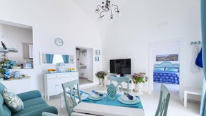 Ein moderner, gut beleuchteter Raum mit einem Esstisch für zwei Personen, einer Couch, einem Fernseher und einem Bett mit blauer und weißer Bettdecke.