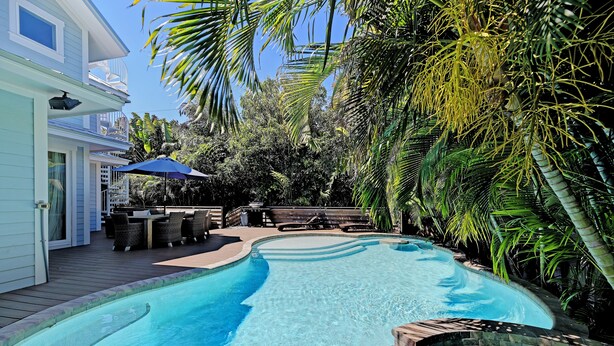 A swimming pool with a wooden deck, surrounded by palm trees and a patio area with outdoor furniture. - Oasis by the Beach managed by the Beach Retreats (Holmes Beach)