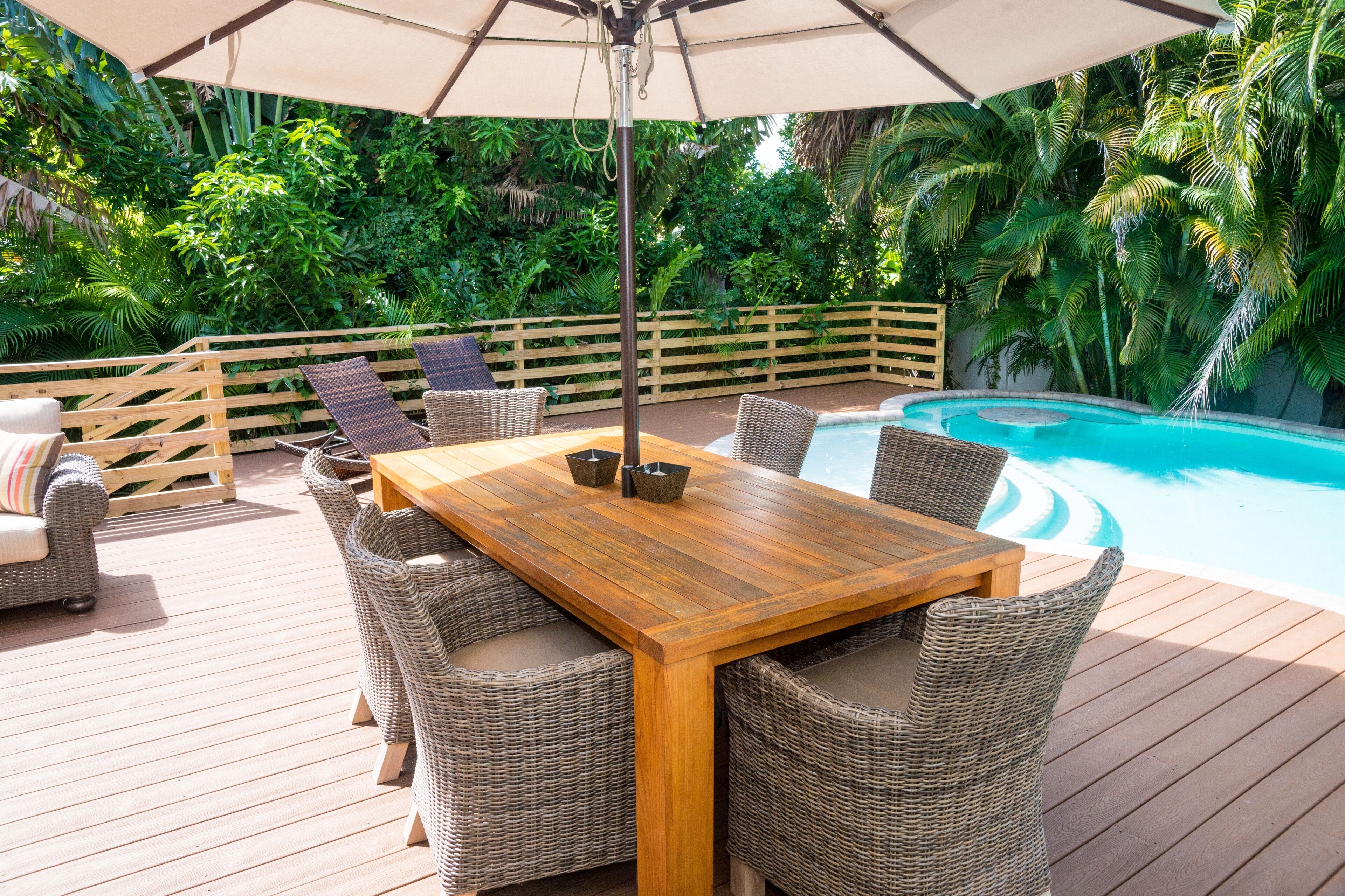 A wooden deck with wicker furniture, a pool, and lush greenery.