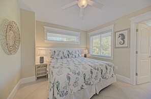 A bedroom with a bed, bedside tables, a ceiling fan, and a window with blinds. - Oasis by the Beach managed by the Beach Retreats (Holmes Beach)