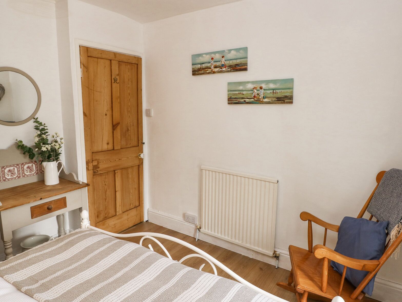 A bedroom with a wooden door, a bed with a striped bedspread, a wooden chair with a blue cushion, and two paintings on the wall.
