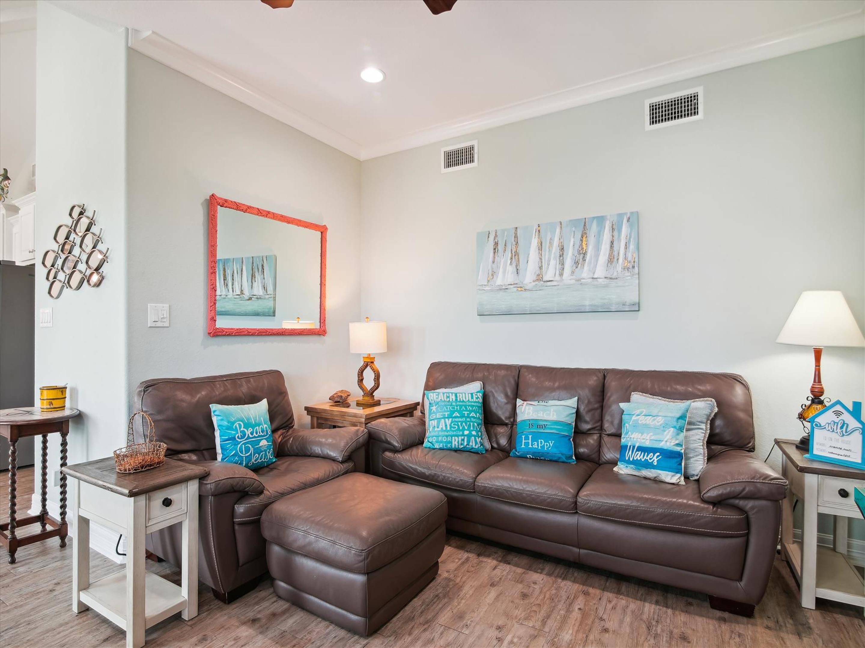 A living room with a brown leather sofa, a small side table, and a lamp. There are wall decorations including a mirror, a painting of sailboats, and a wall-mounted light fixture.