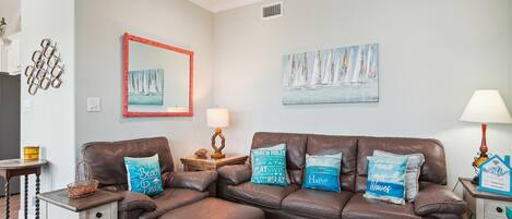 A living room with a brown leather sofa, a small side table, and a lamp. There are wall decorations including a mirror, a painting of sailboats, and a wall-mounted light fixture.