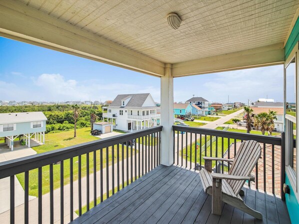 A wooden deck with a railing, a single chair, and a view of a residential area with houses and a road. - Adventure Awaits by Ryson Vacation Rentals (Galveston)