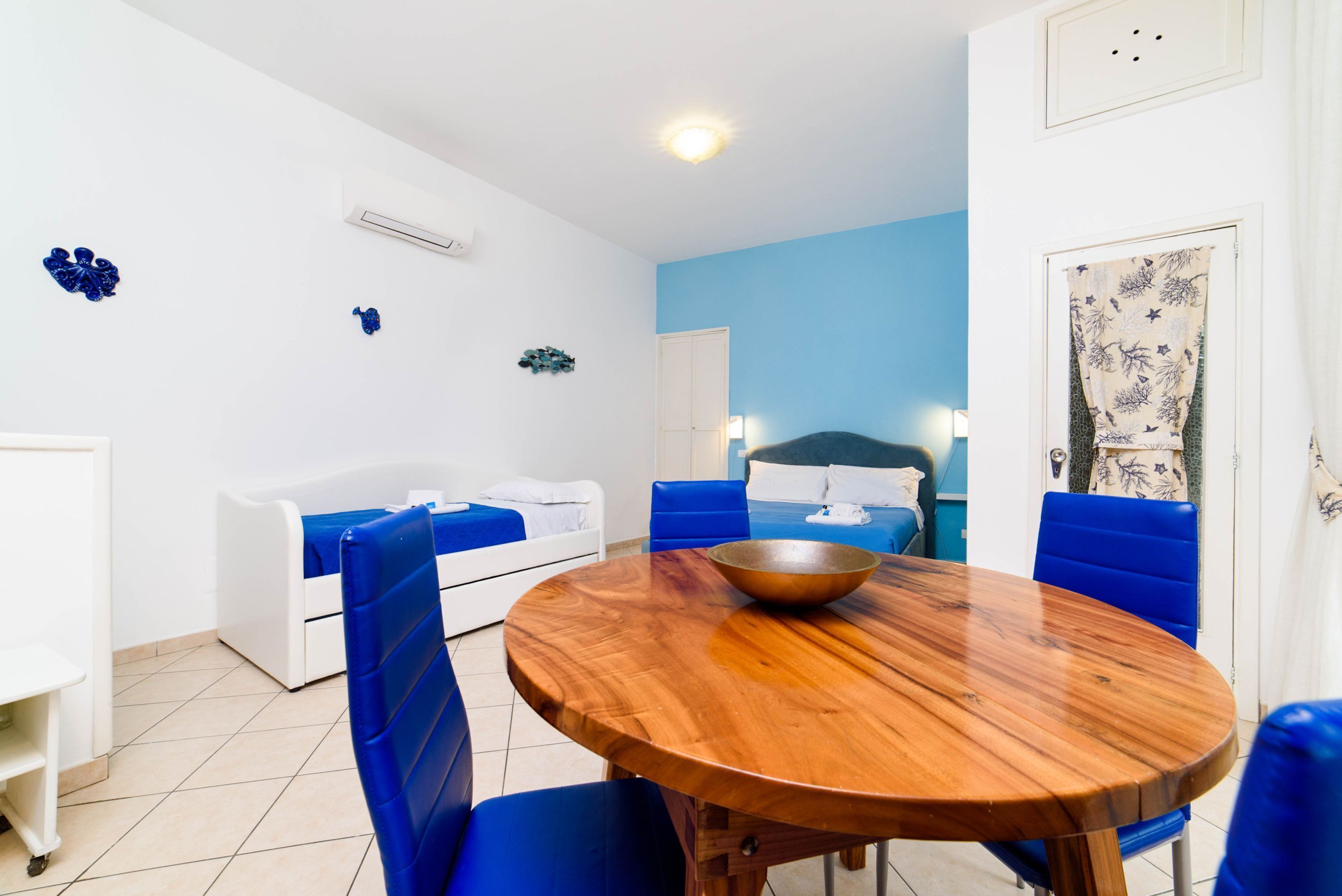 A room with a round wooden table, blue chairs, a white sofa, and a bed with blue cushions.