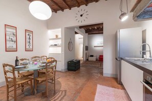 Uma área de refeições com uma mesa redonda posta para quatro pessoas, cadeiras de madeira, um relógio na parede e uma cozinha com geladeira e pia.