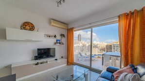 A modern living room with a glass coffee table, a sofa, a flat-screen TV, and a city view through large windows. - Amazing Views of Spinola Bay in Heart of St.julians (St. Julian's)