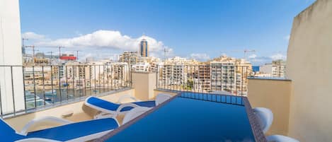 Una piscina en la azotea con sillones de descanso, vista panorámica de la ciudad y cielo despejado.