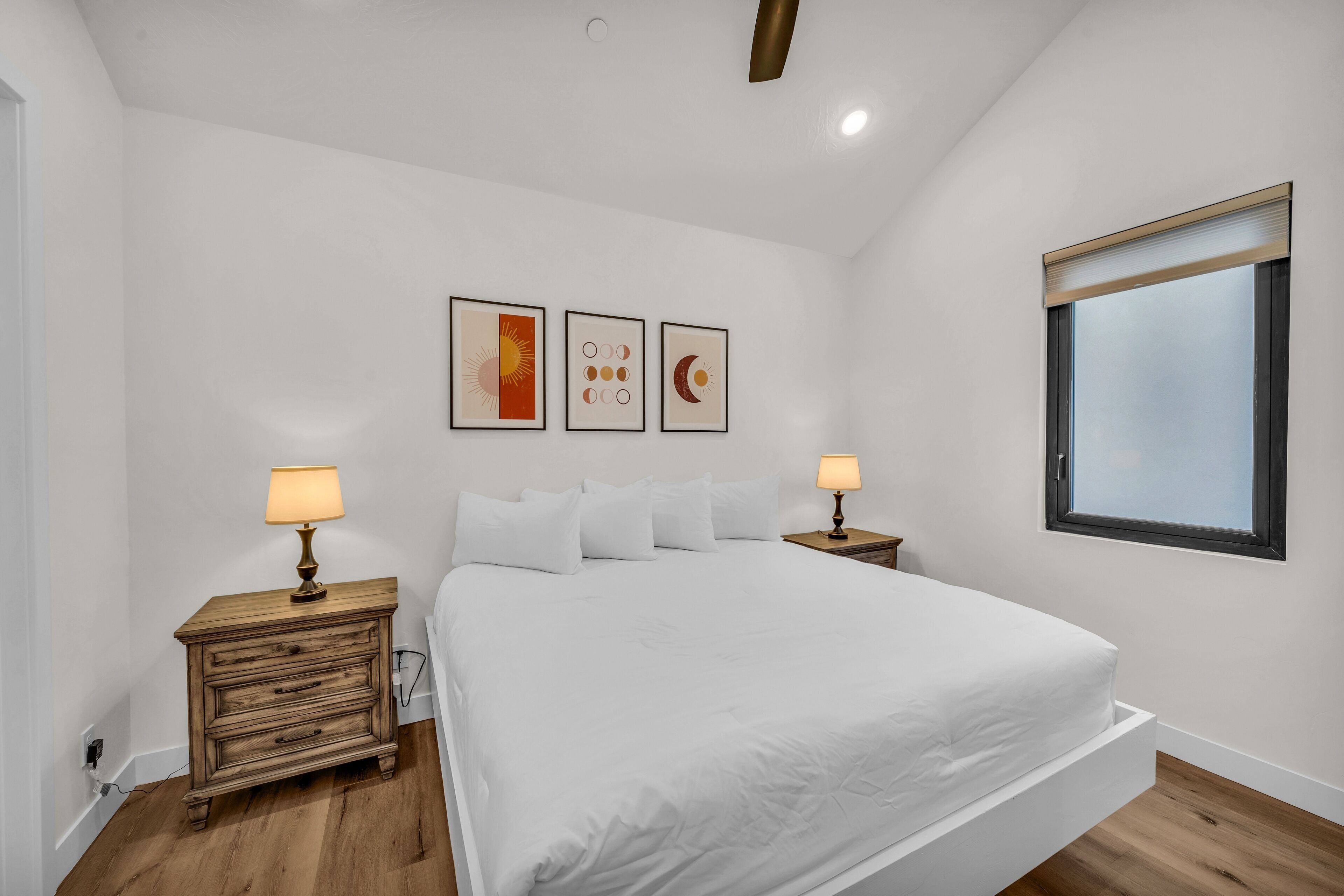 A bedroom with a large bed, two bedside tables with lamps, a window, and three wall-mounted pictures.
