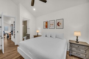A bedroom with a large bed, bedside tables with lamps, a wooden floor, and a bathroom with a shower. - Desert Bungalow #h8 3 Bedroom Home (Hurricane)
