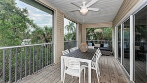 A covered outdoor patio with a table and chairs, a ceiling fan, and a view of palm trees and greenery.