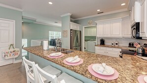 A modern kitchen with granite countertops, white cabinets, and stainless steel appliances. There are bar stools and a sign that reads "VACATION PLEASE".