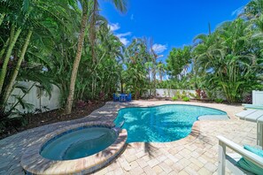 A swimming pool with a hot tub, surrounded by lush tropical plants and a clear blue sky.