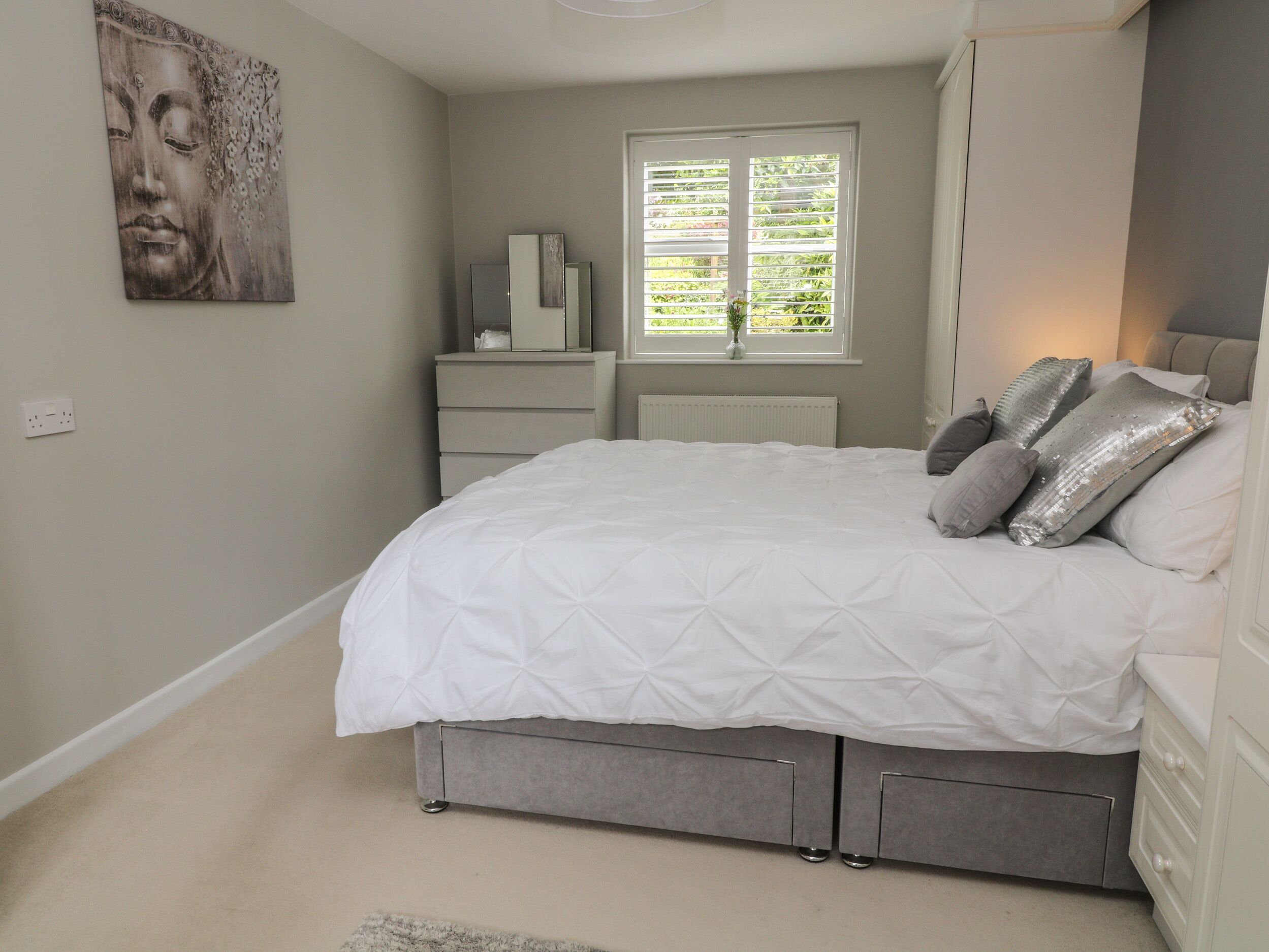 Une chambre à coucher comprenant un lit, des tables de chevet, une commode, une fenêtre avec des stores et une œuvre d’art accrochée au mur.