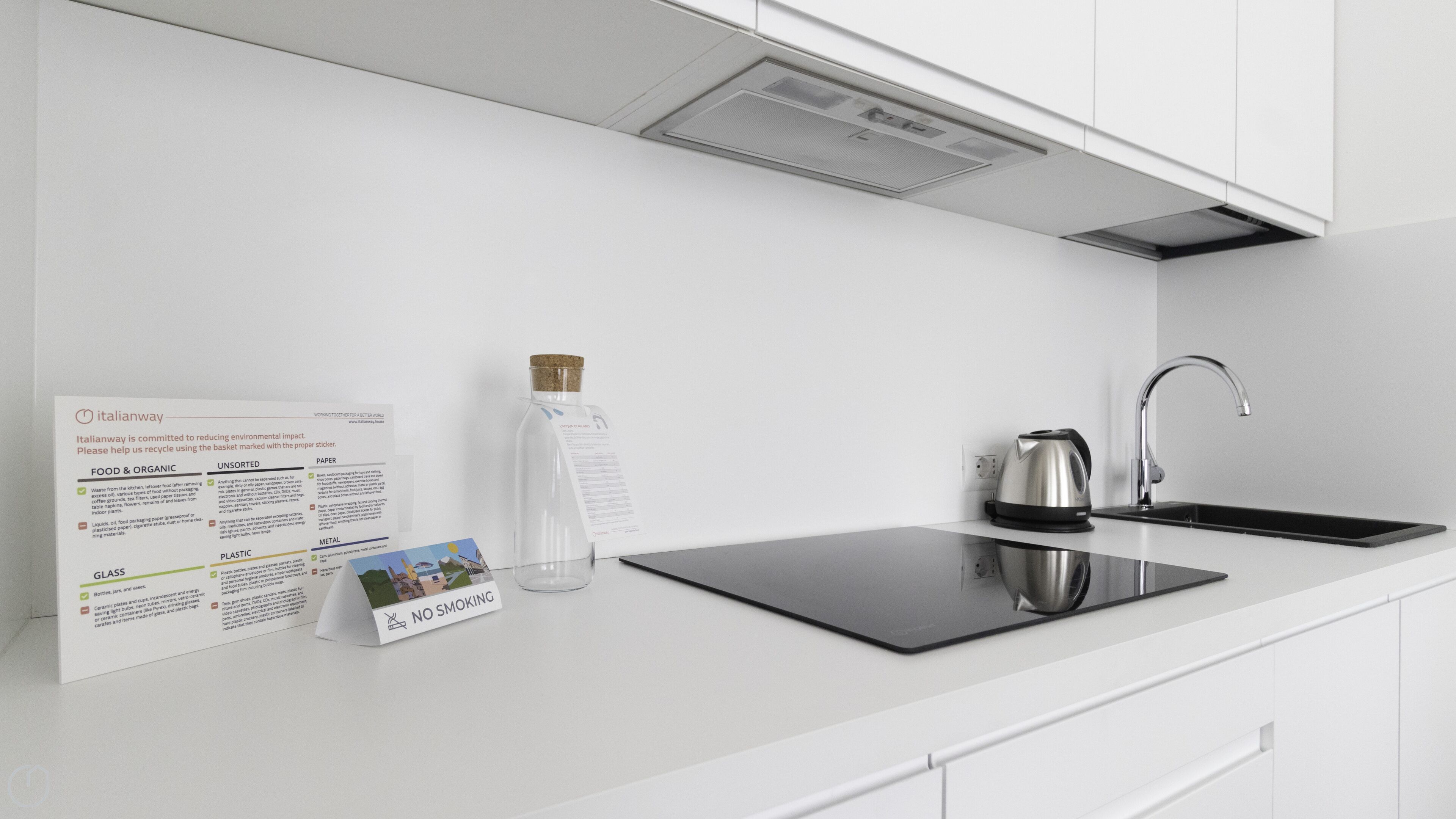 A modern kitchen with white cabinetry, a stainless steel kettle, and a black induction cooktop. A recycling sign is visible on the wall.