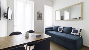 A modern living room with a wooden dining table, two chairs, a blue sofa with patterned cushions, and a large mirror on the wall. - Italianway - Mauro Macchi 65 (Milan)