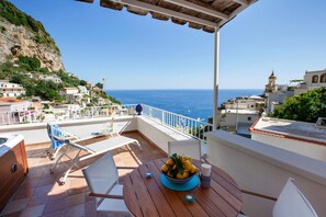 A terrace with a wooden table, chairs, and a bowl of fruit. The terrace overlooks a coastal town with buildings and a cliff. - Medusa Suite in Positano (Positano)