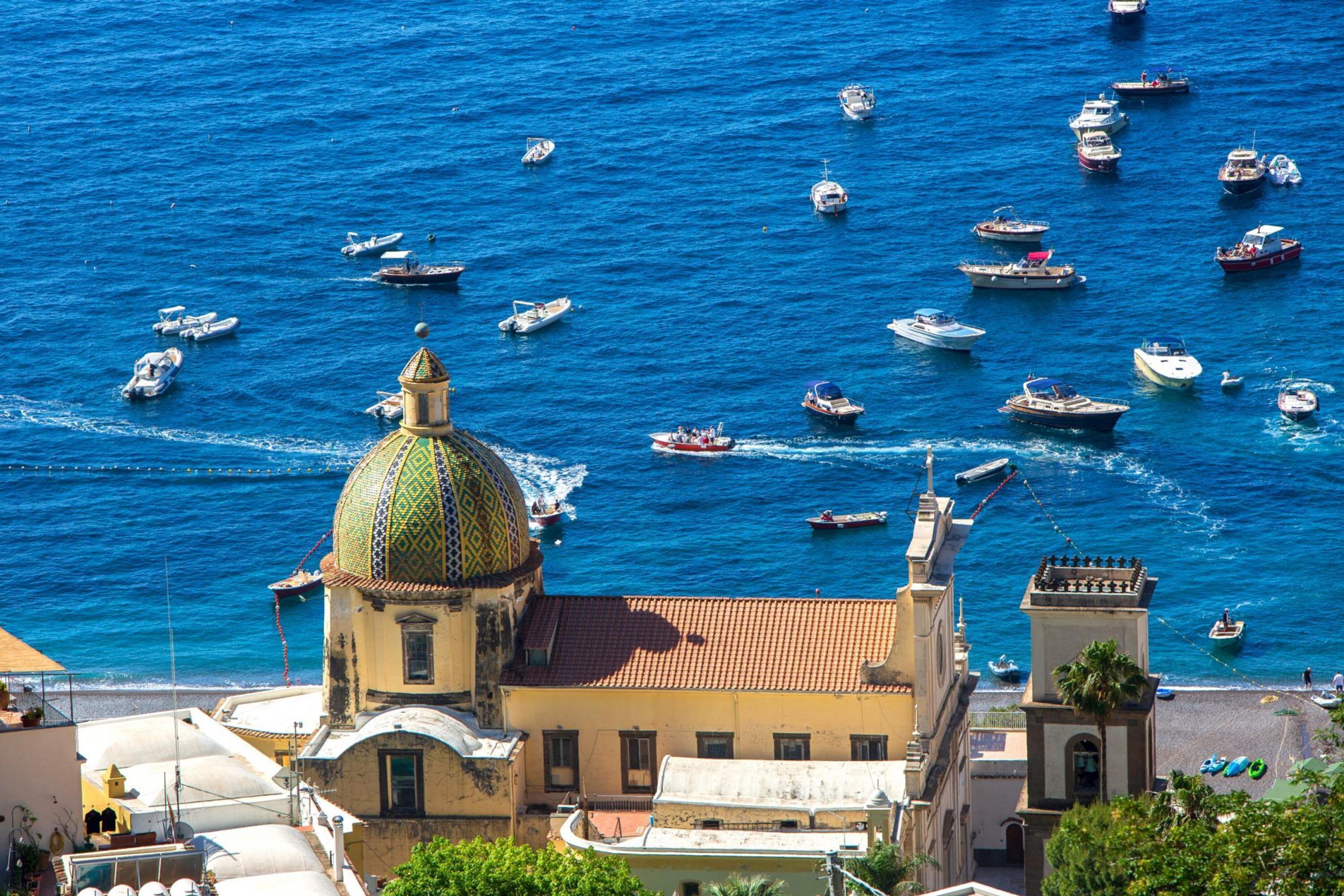A coastal town with a prominent church featuring a green dome, surrounded by numerous boats on the sea.