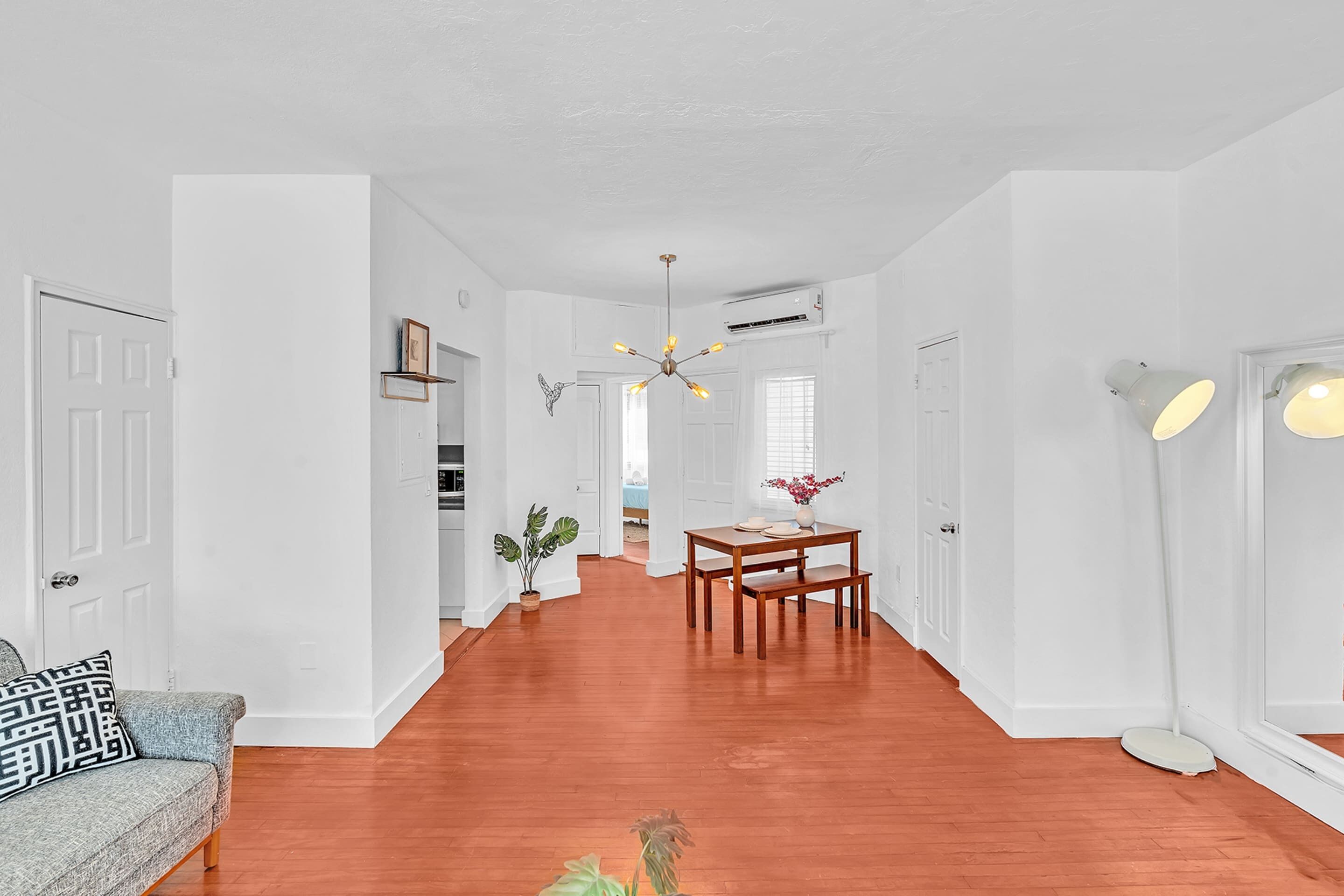 A spacious living area with a wooden floor, white walls, a dining table with chairs, and a ceiling fan.