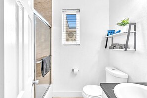 Ein modernes Badezimmer mit Duschabtrennung aus Glas, Toilette, Waschbecken, Fenster mit Jalousien und Ablagefläche für Pflegeprodukte.