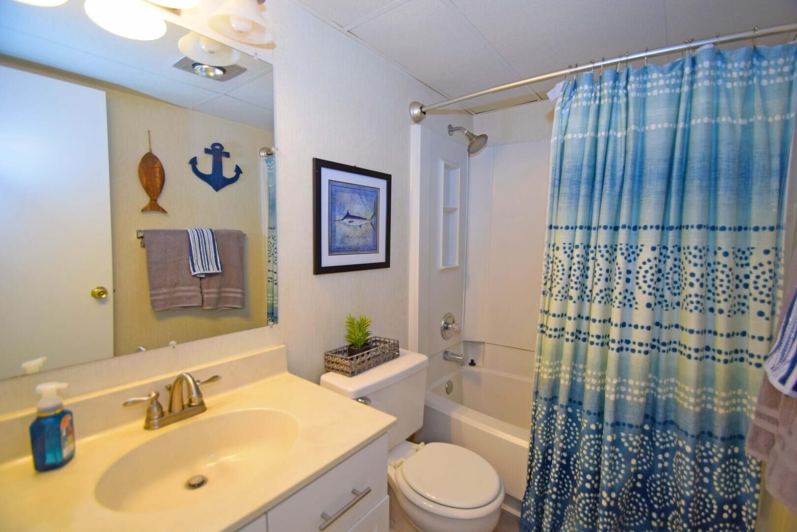 Et badeværelse med vask, toilet og bruser. Der er et indrammet billede på væggen og et håndklæde hængende på døren.