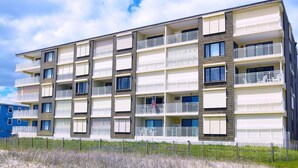 Een meerlaags woongebouw met balkons, een vlag en een zandige voorgrond.