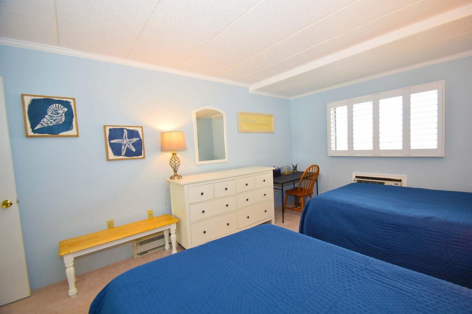 Une chambre avec deux lits, un bureau, une chaise, une commode et une fenêtre avec des stores.
