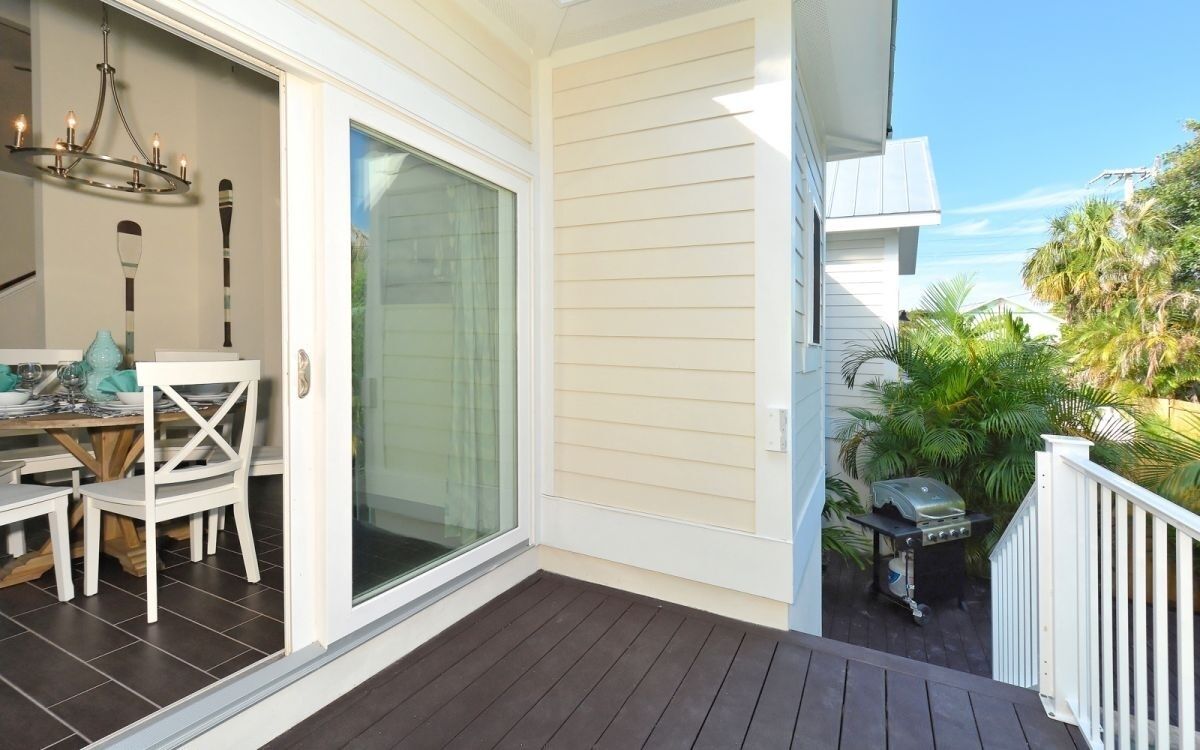 Un'area esterna riparata con una porta scorrevole in vetro che dà su un patio, un tavolo da pranzo con sedie, un barbecue e un piccolo giardino con palme.