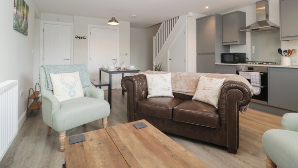 A modern living room with a brown leather sofa, a light blue armchair, and a wooden coffee table. There is a dining table with chairs, a kitchen area with a microwave, and a staircase leading to another level. - The Old School (Ross-on-Wye)