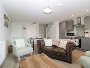 A modern living room with a brown leather sofa, a light blue armchair, and a wooden coffee table. There is a dining table with chairs, a kitchen area with a microwave, and a staircase leading to another level. - The Old School (Ross-on-Wye)