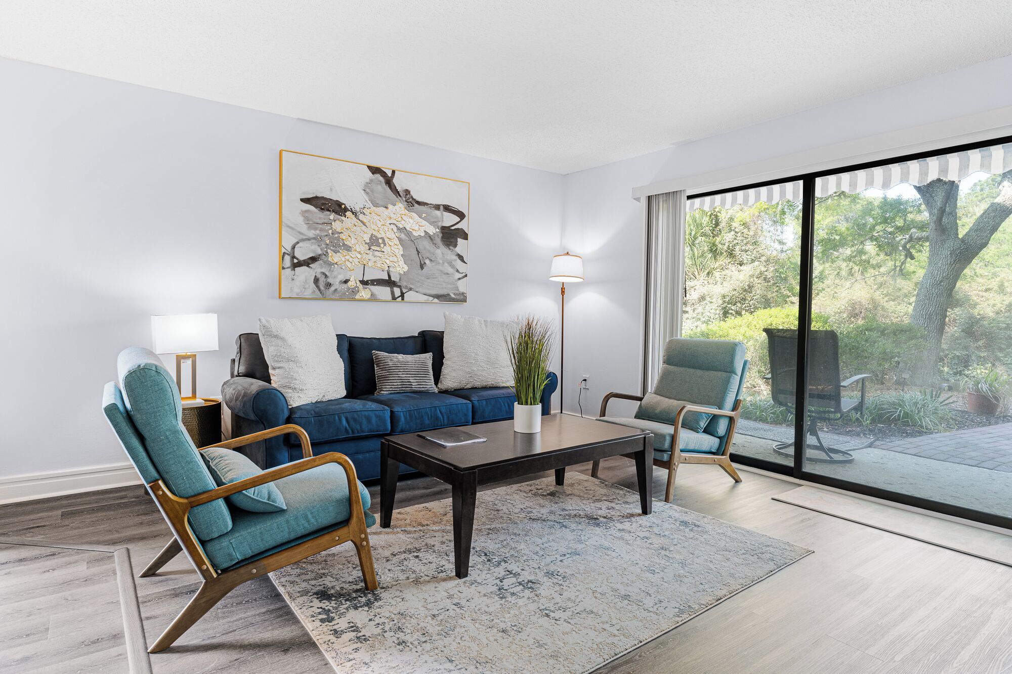 Ein modernes Wohnzimmer mit einem blauen Sofa, zwei Sesseln, einem Couchtisch und einer Stehlampe. Ein großes Fenster bietet Blick auf Grünflächen.