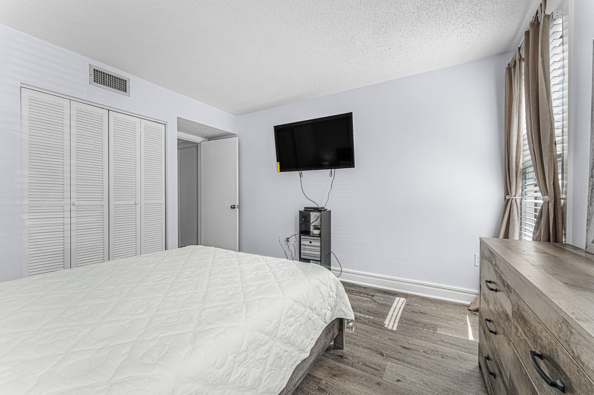 A bedroom with a bed, a TV mounted on the wall, a dresser, and a closet with sliding doors.