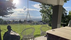 A patio with a swing chair and a table, overlooking a grassy area and a distant view of houses and mountains. - Sunset Views! Indoor Sport Court, Pool table, Golf Course, 12 min to downtown  (North Salt Lake)
