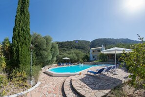 A swimming pool with lounge chairs and umbrellas, surrounded by stone tiles and greenery. - Apartment Ilex - Agios Nikitas Villas (Lefkada)