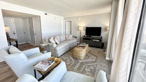 Ein modernes Wohnzimmer mit einer Couch, Sesseln, einem Couchtisch und einem Fernseher. Im Hintergrund sind Türen und eine Lampe zu sehen.