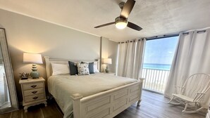 Ein Schlafzimmer mit einem großen Bett, zwei Nachttischen mit Lampen, einem Deckenventilator und Blick auf den Strand durch einen Balkon.