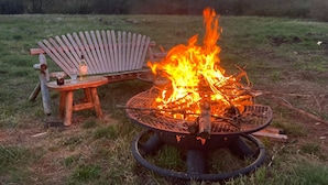 A campfire with a metal grill, a wooden Adirondack chair, and a small table with a bottle and glass. - Cozy Modern Cabin Surrounded by Nature & Pet Friendly! (Grand Lake)