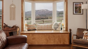 A cozy seating area with a view of mountains through a bay window, a leather couch, a wooden bench, and a rustic decor. - Cozy Modern Cabin Surrounded by Nature & Pet Friendly! (Grand Lake)