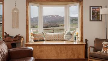 A cozy seating area with a view of mountains through a bay window, a leather couch, a wooden bench, and a rustic decor.