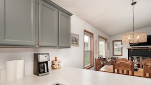 A kitchen with grey cabinets, a coffee maker, and a dining area with wooden chairs and a table. - Cozy Modern Cabin Surrounded by Nature & Pet Friendly! (Grand Lake)