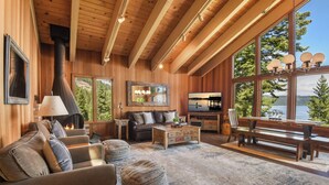 A cozy cabin interior with a fireplace, wooden furniture, a large TV, and a view of the outdoors. - Eagles Perch Cabin - Pano Lake Views, Fido Friendly (Carnelian Bay)