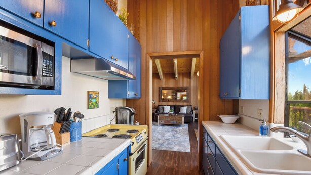 A kitchen with blue cabinets, a microwave, a stove, and a coffee maker. There is a window with a view of trees. - Eagles Perch Cabin - Pano Lake Views, Fido Friendly (Carnelian Bay)