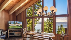A wooden interior with a large window overlooking a lake, a dining table, and a television on a wooden stand. - Eagles Perch Cabin - Pano Lake Views, Fido Friendly (Carnelian Bay)