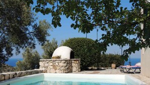 Ein Swimmingpool mit Steinmauer und Pizzaofen, umgeben von Bäumen und Blick auf das Meer.