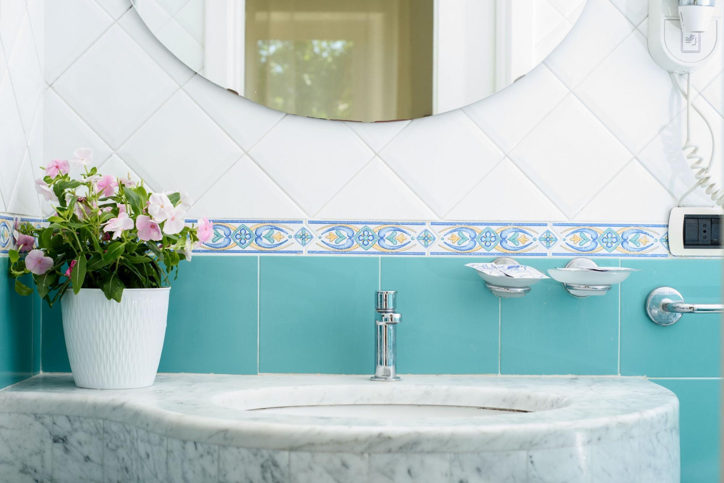 Une salle de bain avec un lavabo en marbre blanc, une plante en pot aux fleurs roses, un miroir et un mur recouvert de carreaux bleus.