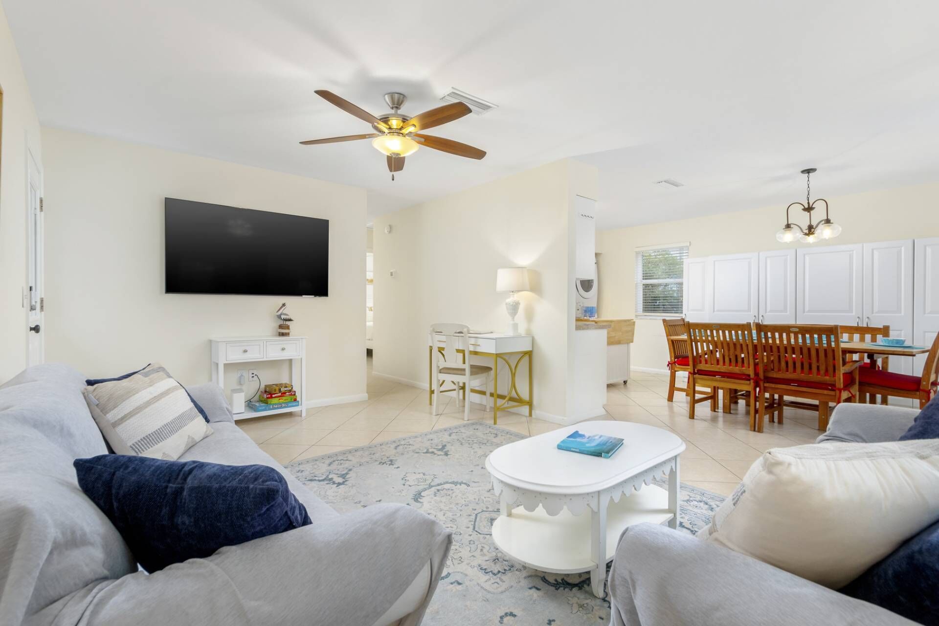 A living room with a flat-screen TV, a ceiling fan, a dining area with wooden chairs and a white table, and a kitchen area with white cabinets.